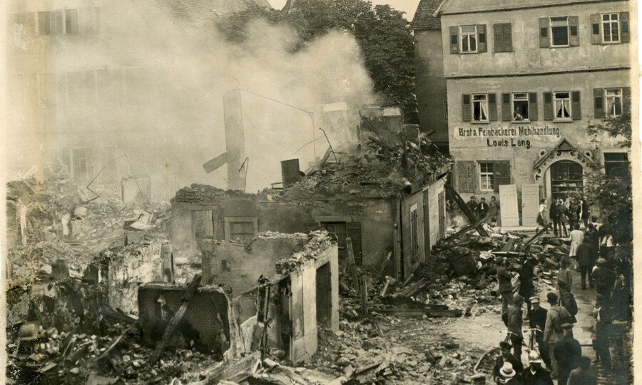 Schwarz-Weiß-Fotografie des Stadtbrands von Bietigheim im Jahr 1921: Zerstörte und eingestürzte Gebäude rauchen noch, Trümmer bedecken die Straße. Zahlreiche Menschen stehen am Rand der Brandruine. Im Hintergrund ist unter anderem die Feinbäckerei Louis Leng (das heutige Stadtmuseum Hornmoldhaus) zu sehen.