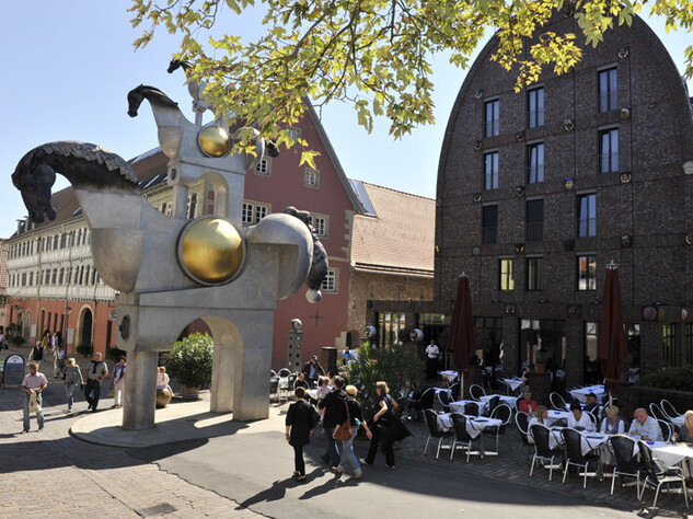 Großer Turm aus Pferdeskulpturen vor einem Backsteinhaus mit rundem Dach