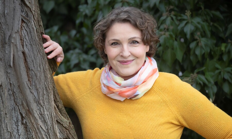 Bild der Autorin Nina Blazon, die an einen Baum gelehnt ist mit gelbem Pulli und Schal