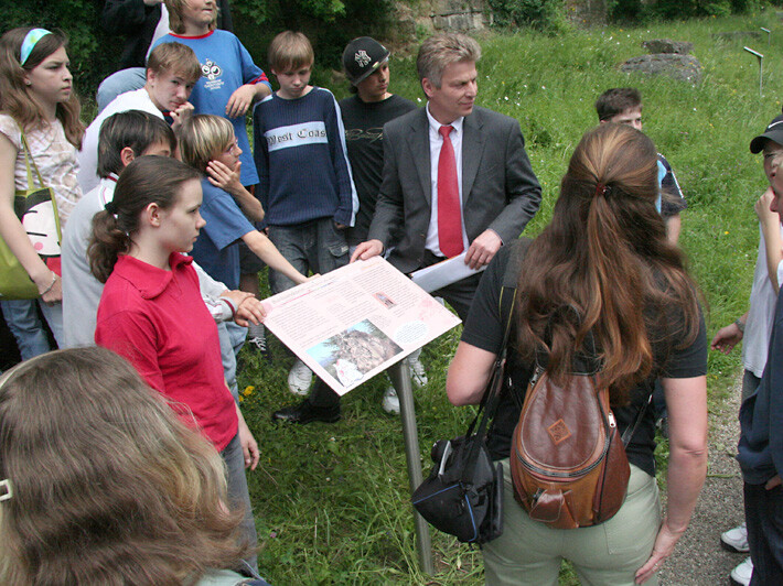 Schüler stehen um eine Lehrtafel auf einem Pfad und hören einer Lehrerin zu.