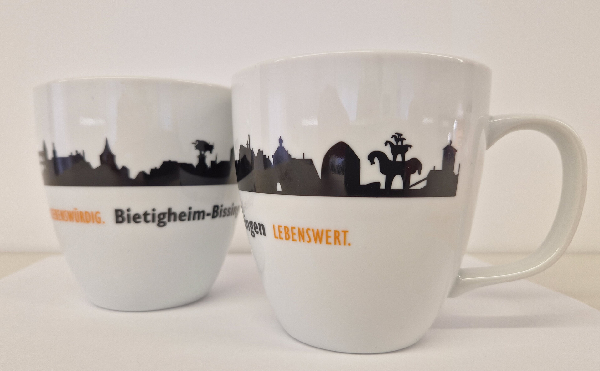 Tasse (7,50 €) Zwei weiße Tassen stehen vor weißem Hintergrund. Auf beiden Tassen ist eine schwarze Stadtsilhouette und Name der Stadt zu sehen.